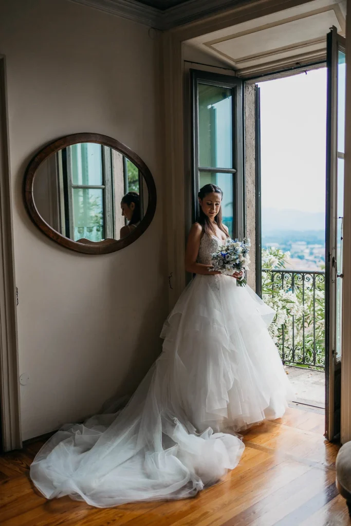 Dream Wedding at Villa Le Due Torrette in Erba: The Elegance of a Lake Como Celebration 26 Bride standing by the window at Villa Le Due Torrette, looking out at Lake Como