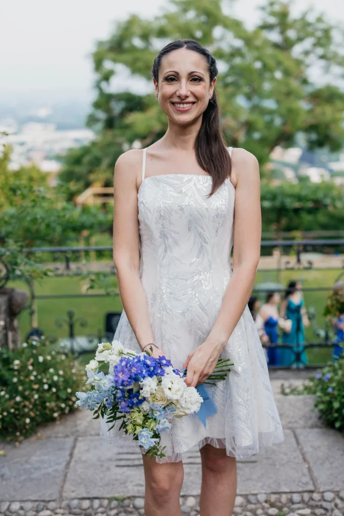 Dream Wedding at Villa Le Due Torrette in Erba: The Elegance of a Lake Como Celebration 88 Bridesmaid holding bouquet of blue and white flowers in outdoor ceremony setting