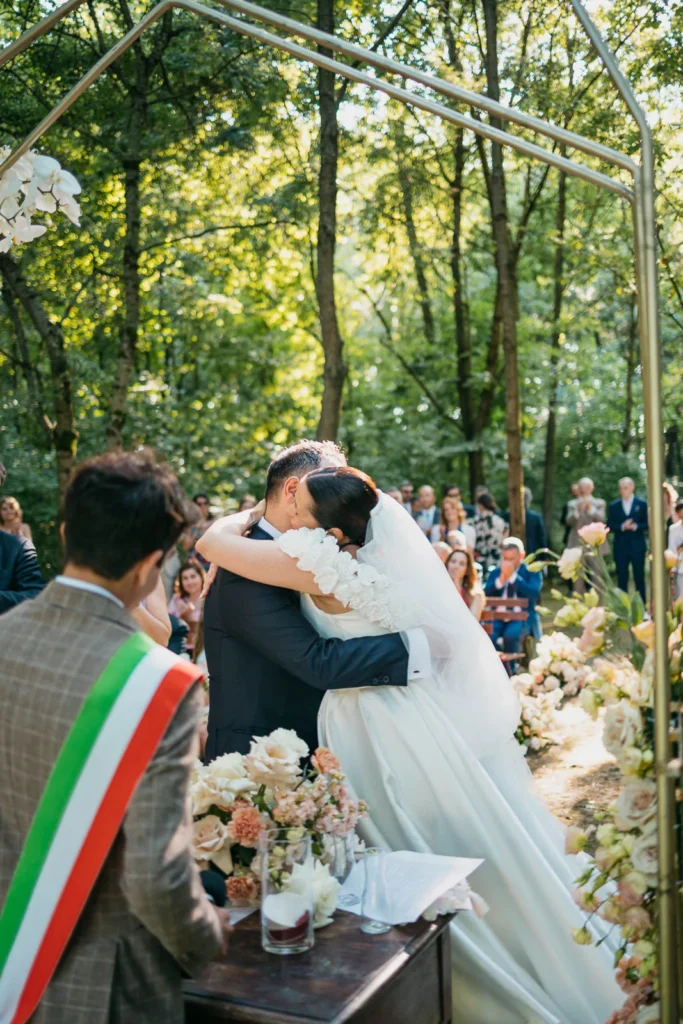 First kiss of bride and groom surrounded by trees and guests